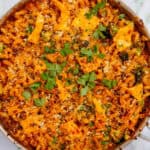 A pan of baked mince pasta with melted cheese, broccoli, and herbs on top, garnished with fresh parsley. A small dish with more parsley sits nearby on a white surface.