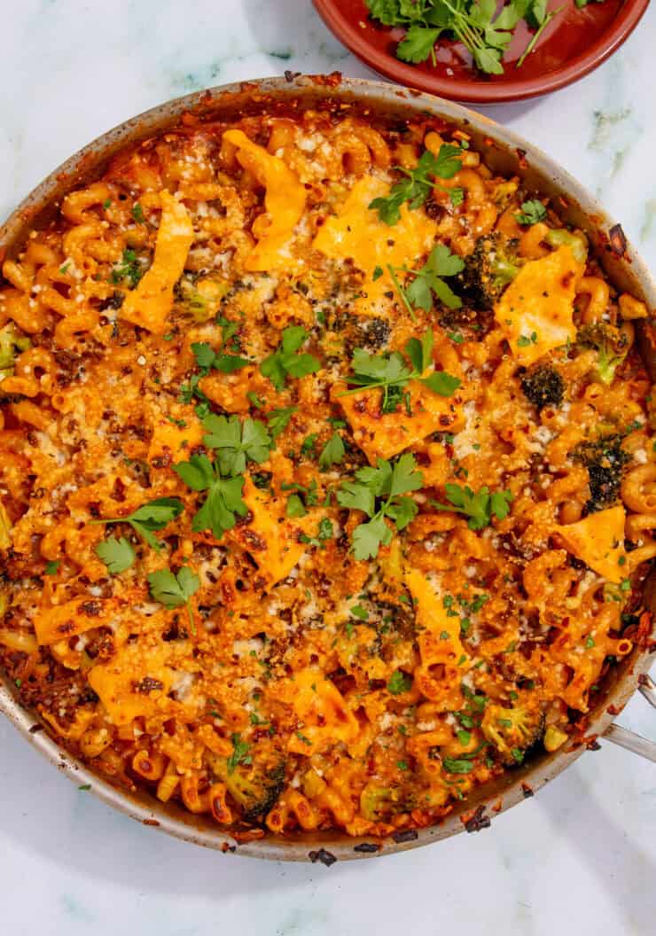 A pan of baked mince pasta with melted cheese, broccoli, and herbs on top, garnished with fresh parsley. A small dish with more parsley sits nearby on a white surface.