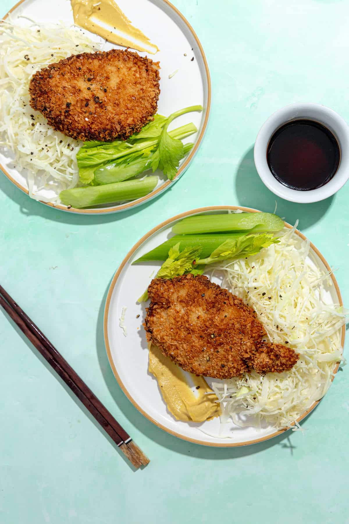 Two plates with breaded, fried cutlets, shredded cabbage, mustard, celery sticks, and roasted corn. A small bowl of dark dipping sauce and a pair of chopsticks rest on a light green surface beside the plates.