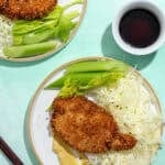 Two plates with breaded, fried cutlets, shredded cabbage, celery sticks, next to a bowl of dark dipping sauce and a pair of chopsticks on a light turquoise background.