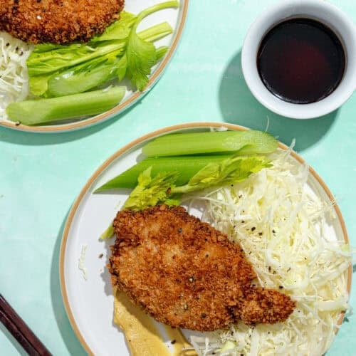 Two plates with breaded, fried cutlets, shredded cabbage, celery sticks, next to a bowl of dark dipping sauce and a pair of chopsticks on a light turquoise background.