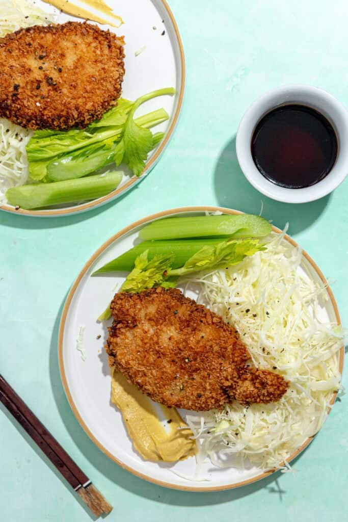 Two plates with breaded, fried cutlets, shredded cabbage, celery sticks, next to a bowl of dark dipping sauce and a pair of chopsticks on a light turquoise background.