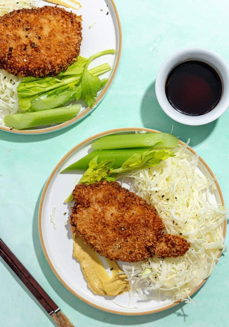 Two plates with breaded, fried cutlets, shredded cabbage, celery sticks, next to a bowl of dark dipping sauce and a pair of chopsticks on a light turquoise background.