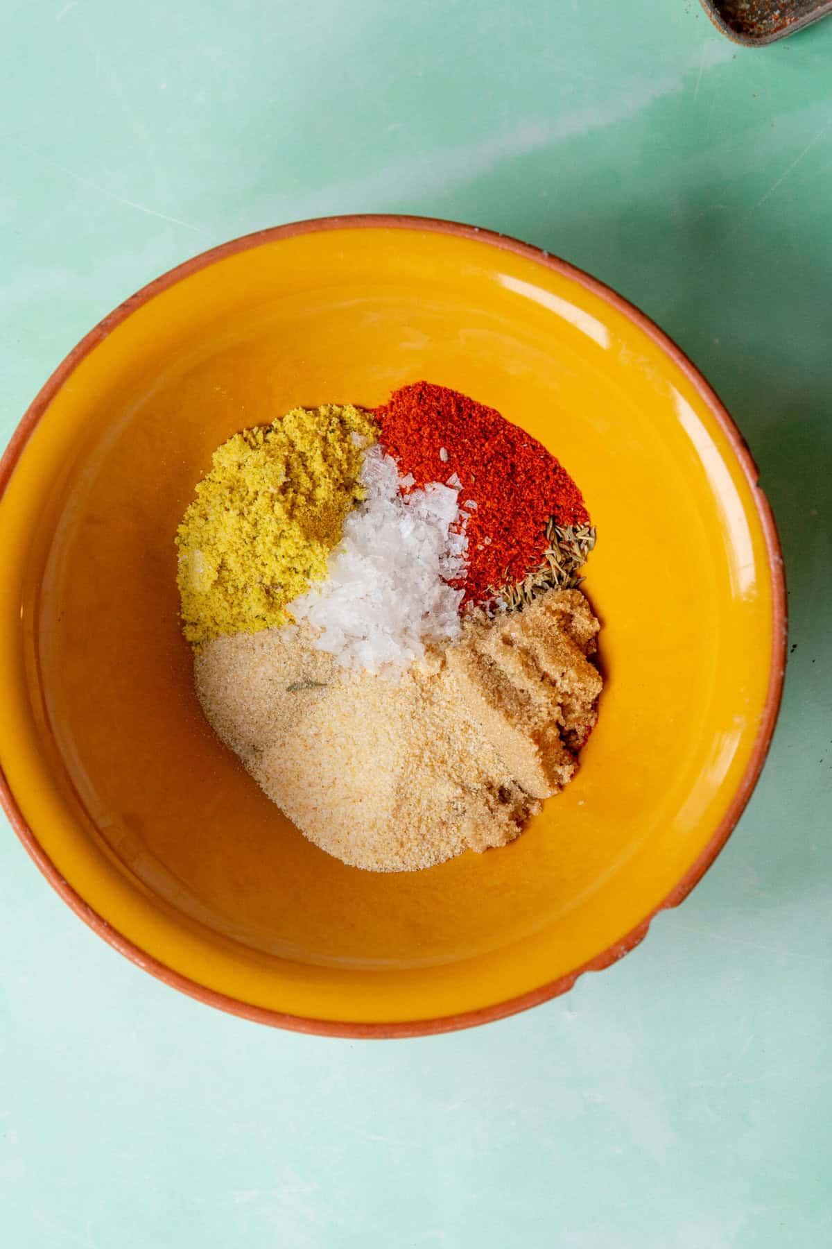 A yellow bowl contains piles of various spices and seasonings, including red chili powder, collard greens seasoning, white flakes, and beige granules, arranged separately on a light green surface.