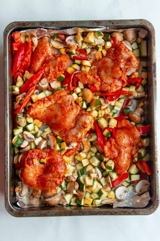 A baking sheet topped with four pieces of seasoned chicken thighs surrounded by chopped zucchini, red bell peppers, mushrooms, and a dense bean salad, ready to be cooked.