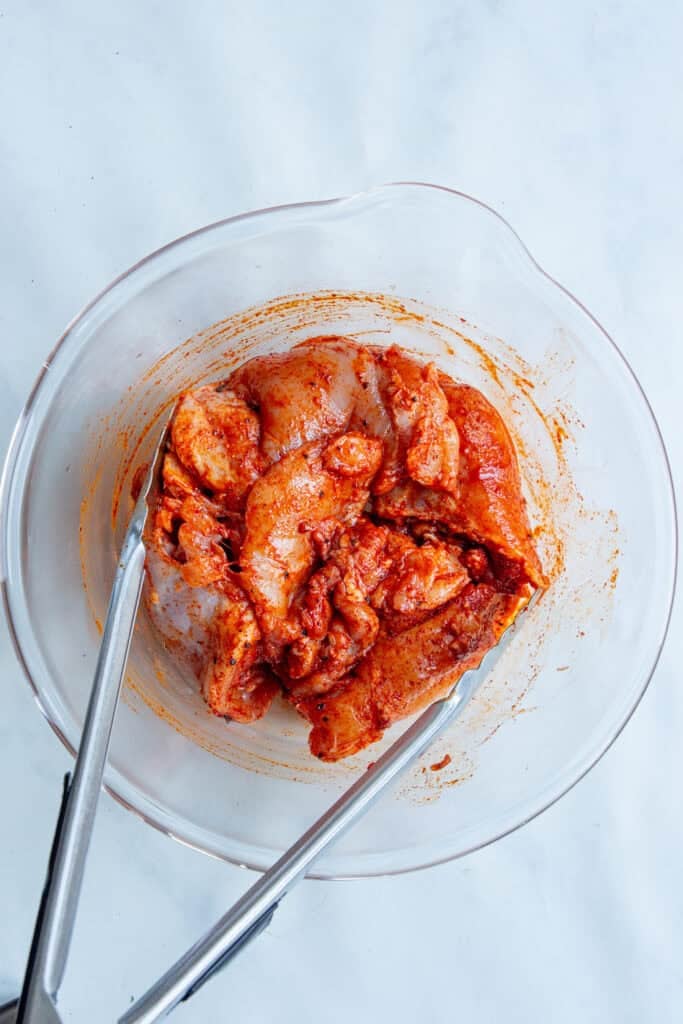 Raw chicken pieces coated in red marinade sit in a clear glass bowl with metal tongs on a white surface, ready to be paired with a dense bean salad for a hearty meal.