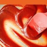 A pink spatula stirs thick, glossy red low sugar BBQ sauce in a metal bowl. Text above reads: In less than 10 minutes. Low Sugar BBQ Sauce.