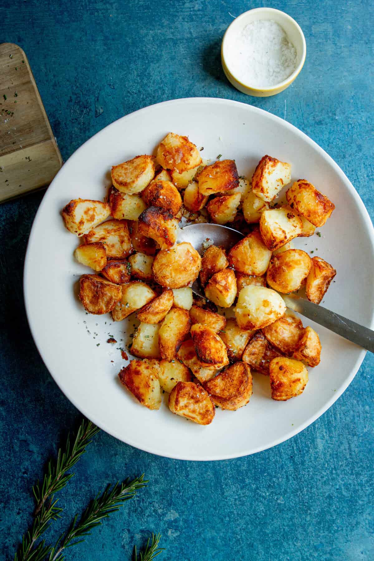 A white plate filled with golden, crispy roasted potatoes sits on a blue surface, with a serving spoon resting on the plate. A small bowl of salt, a sprig of rosemary, and a wooden board are nearby.