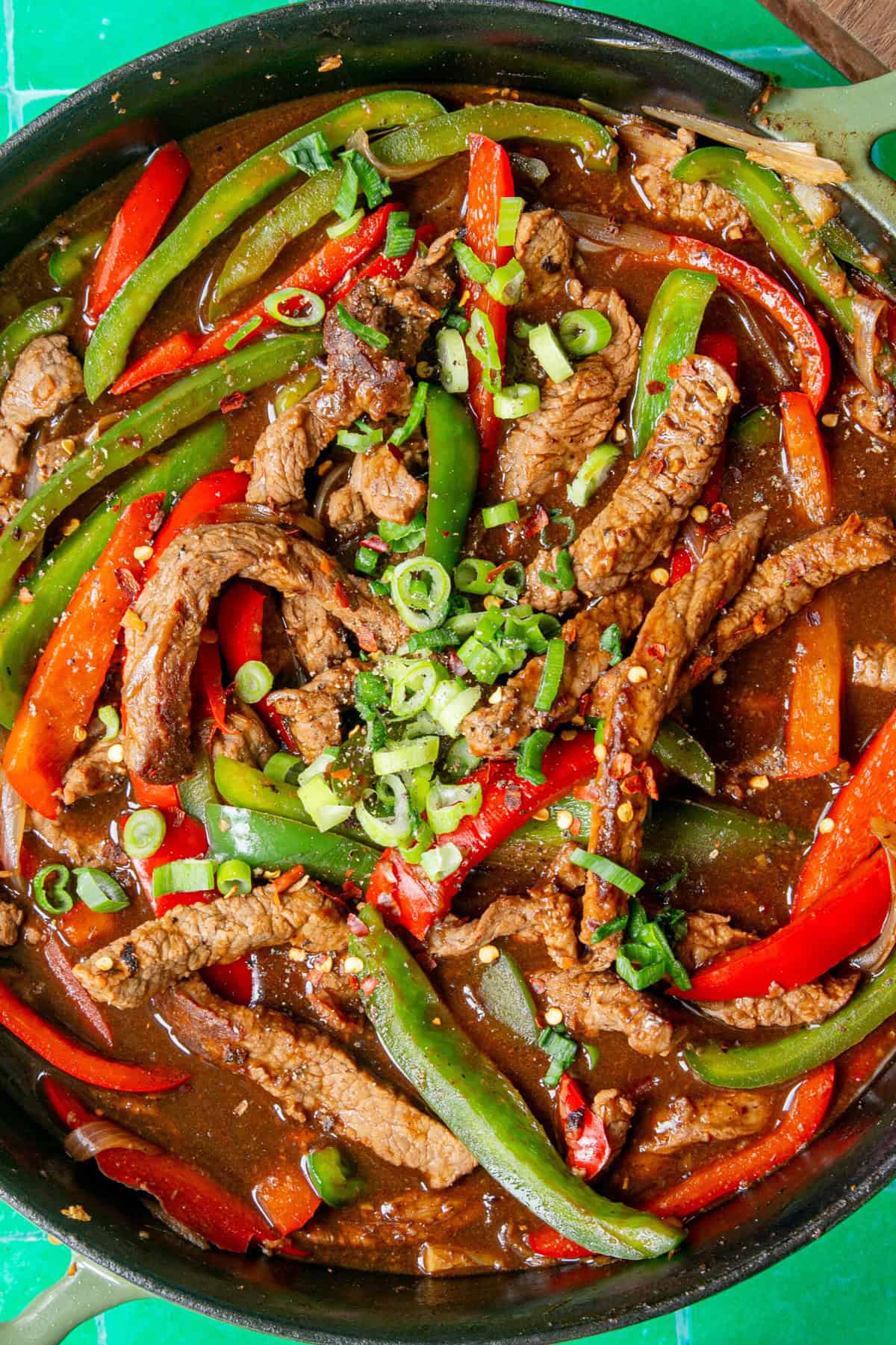 A skillet filled with beef stir-fry, featuring sliced beef, red and green bell peppers, chopped green onions, and sweet roasted corn in a rich brown sauce.