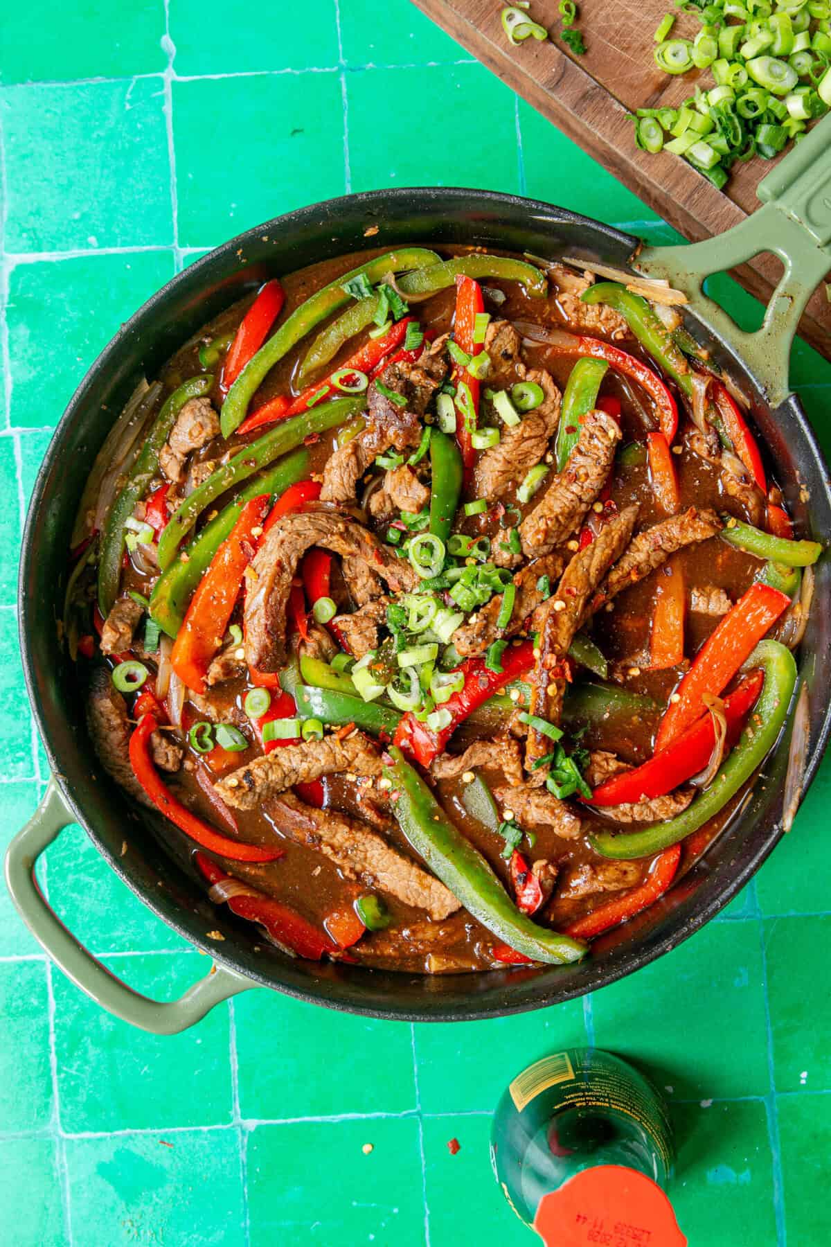 A skillet filled with sliced beef, red and green bell peppers, onions, roasted corn, and sauce, garnished with sliced green onions, sits on a bright green tiled surface next to a wooden board with more chopped green onions.