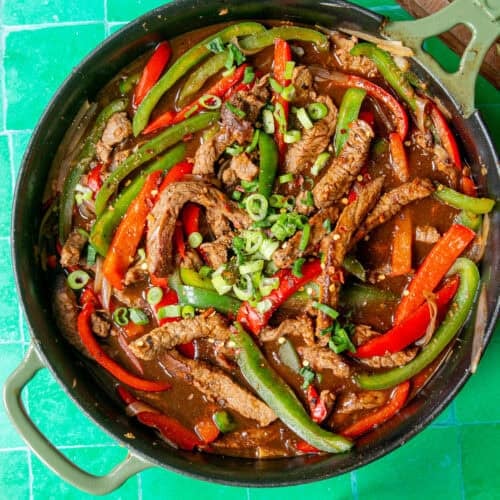 A skillet filled with sliced beef, red and green bell peppers, onions, and sauce, garnished with chopped green onions, sits on a bright green tiled surface.
