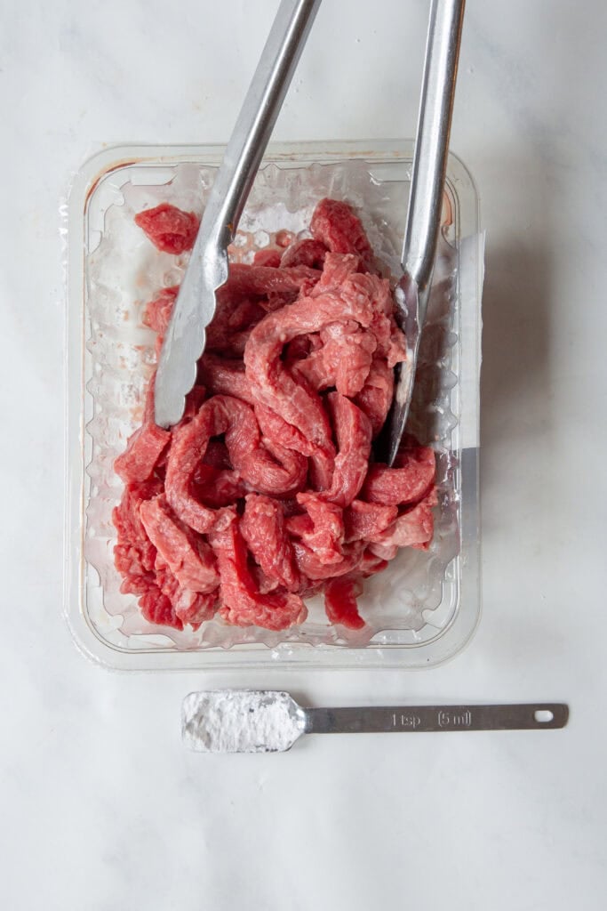 A pair of metal tongs holding strips of raw beef in a plastic tray, with a measuring spoon containing white powder, labeled 1 tsp (5 mL), placed below the tray on a white surface beside some roasted corn.
