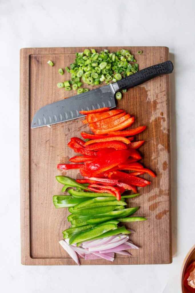 A wooden cutting board with sliced green onions, red bell peppers, green bell peppers, roasted corn, and red onions, with a black-handled chef's knife resting on top.