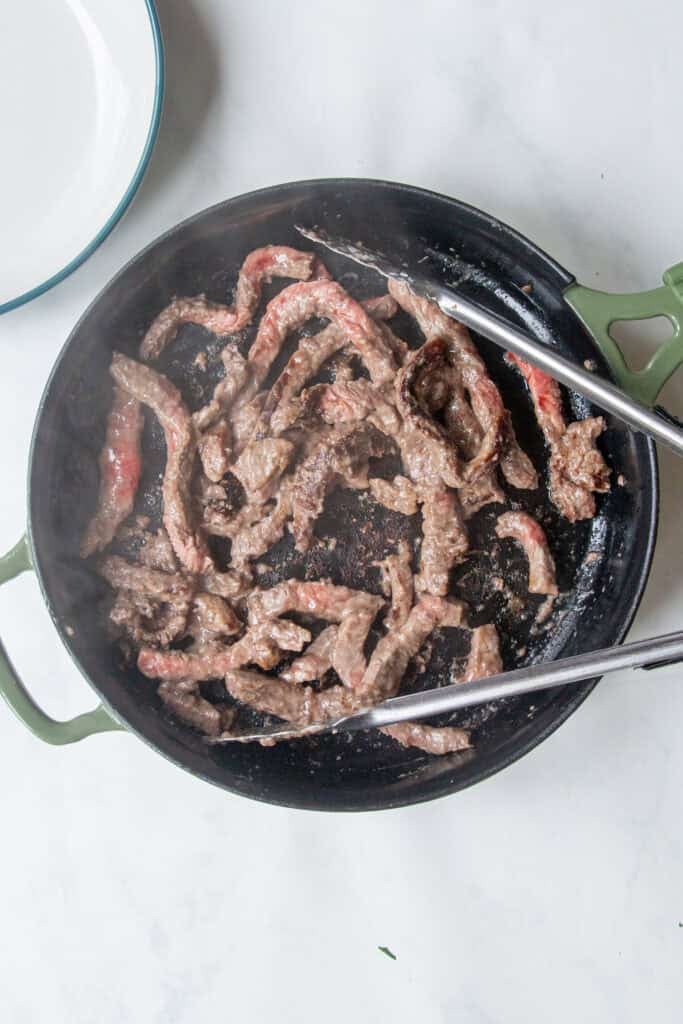 Sliced beef strips, red bell pepper pieces, and roasted corn are being cooked in a round, green-handled skillet with metal tongs on a white surface. Steam is visible, indicating the food is hot.