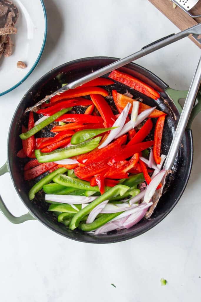 A cast iron skillet filled with sliced red and green bell peppers, onions, and roasted corn sits on a white countertop, with metal tongs resting on the side.