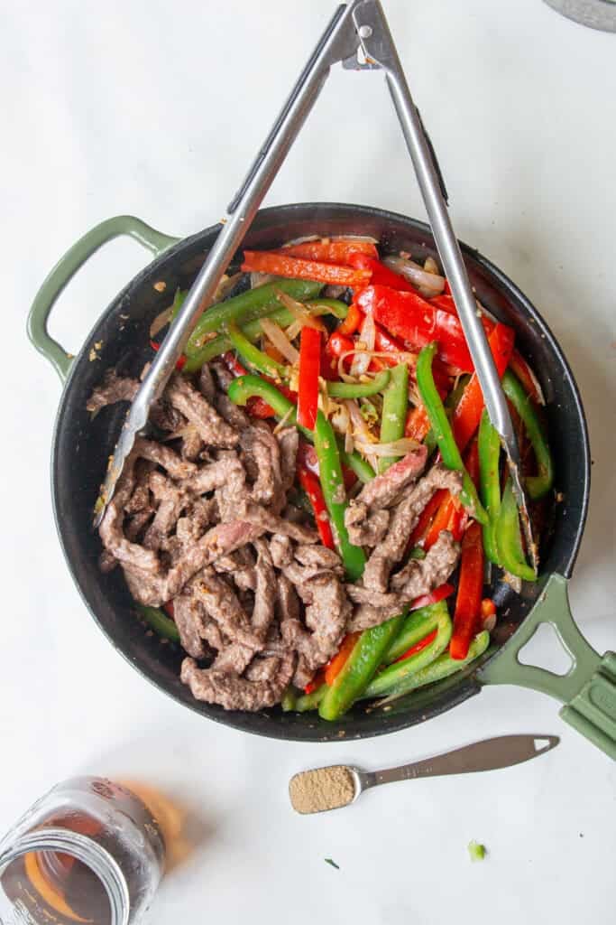 A skillet filled with cooked beef strips, sliced red and green bell peppers, onions, and roasted corn, with metal tongs resting on top. Nearby are a measuring spoon with spices and a jar containing liquid.
