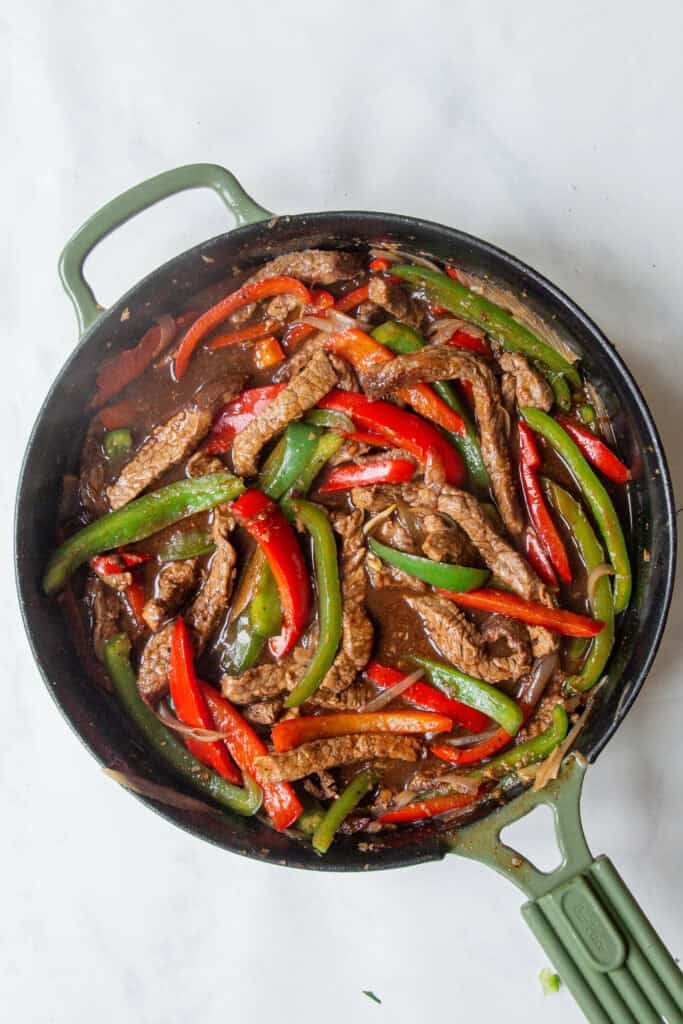 A skillet filled with cooked beef strips, red and green bell peppers, roasted corn, and sauce, placed on a white surface.