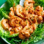 A plate of lettuce leaves topped with Boom Boom Shrimp coated in a creamy orange sauce, garnished with chopped herbs and crispy breadcrumbs, served on a green tiled surface.