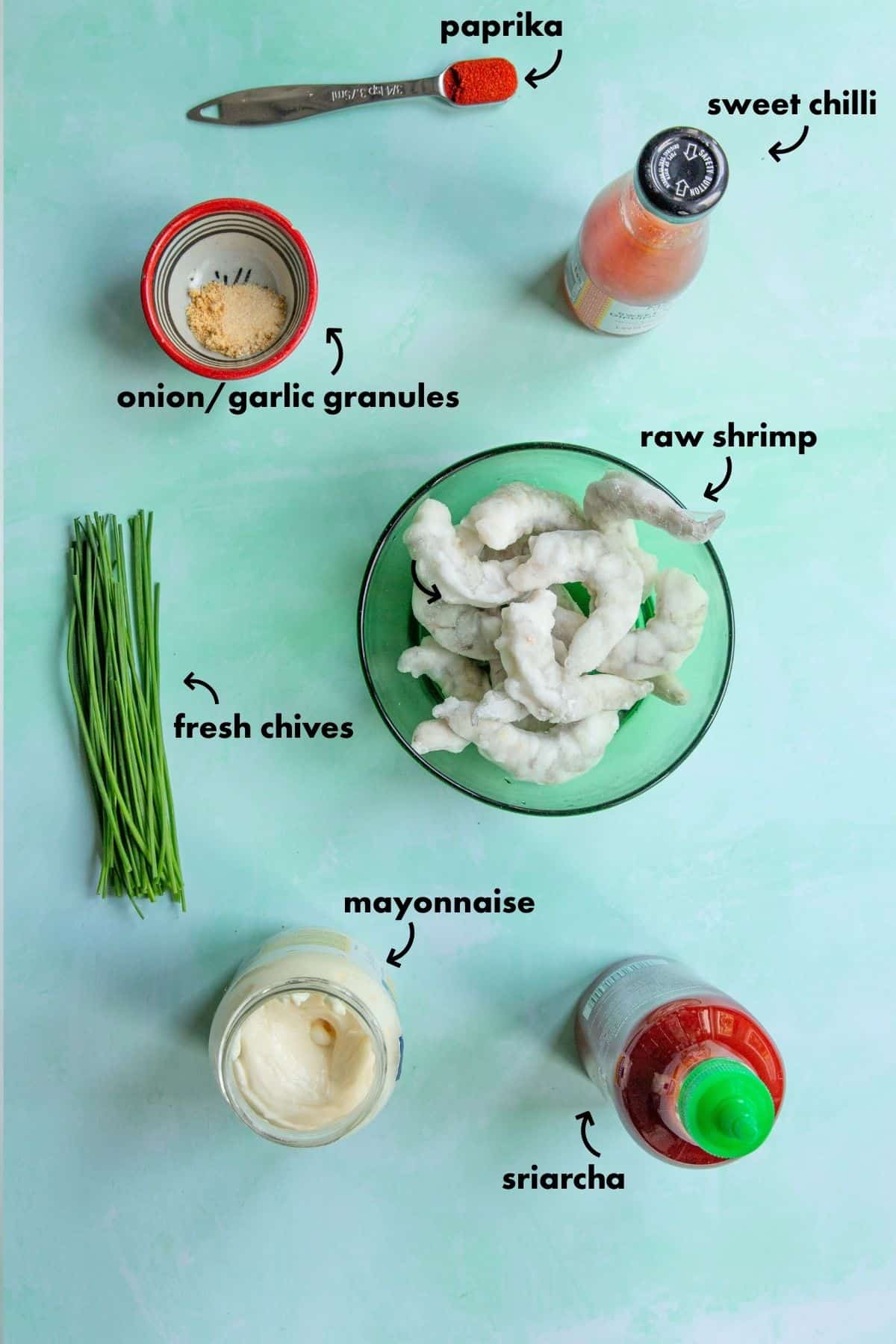 A flat lay of ingredients for Boom Boom Shrimp on a light green surface: a bowl of raw shrimp, fresh chives, mayonnaise, sweet chili sauce, sriracha, onion/garlic granules, and a teaspoon of paprika, each labeled with text.