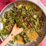 A skillet with cooked collard greens and diced bacon in flavorful collard greens seasoning broth, stirred with a wooden spatula, placed on a pink tiled surface with a yellow bowl and a white cloth nearby.