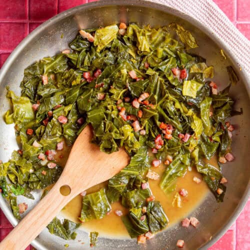 A skillet with cooked collard greens and diced bacon in flavorful collard greens seasoning broth, stirred with a wooden spatula, placed on a pink tiled surface with a yellow bowl and a white cloth nearby.