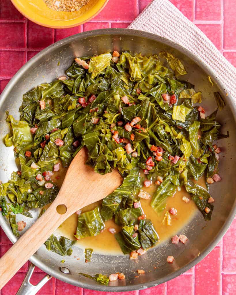 A skillet with cooked collard greens and diced bacon in flavorful collard greens seasoning broth, stirred with a wooden spatula, placed on a pink tiled surface with a yellow bowl and a white cloth nearby.