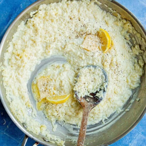 A skillet of creamy rice risotto, garnished with cracked black pepper, two lemon wedges, and a pat of butter, with a wooden spoon resting inside the pan on a blue tiled surface.
