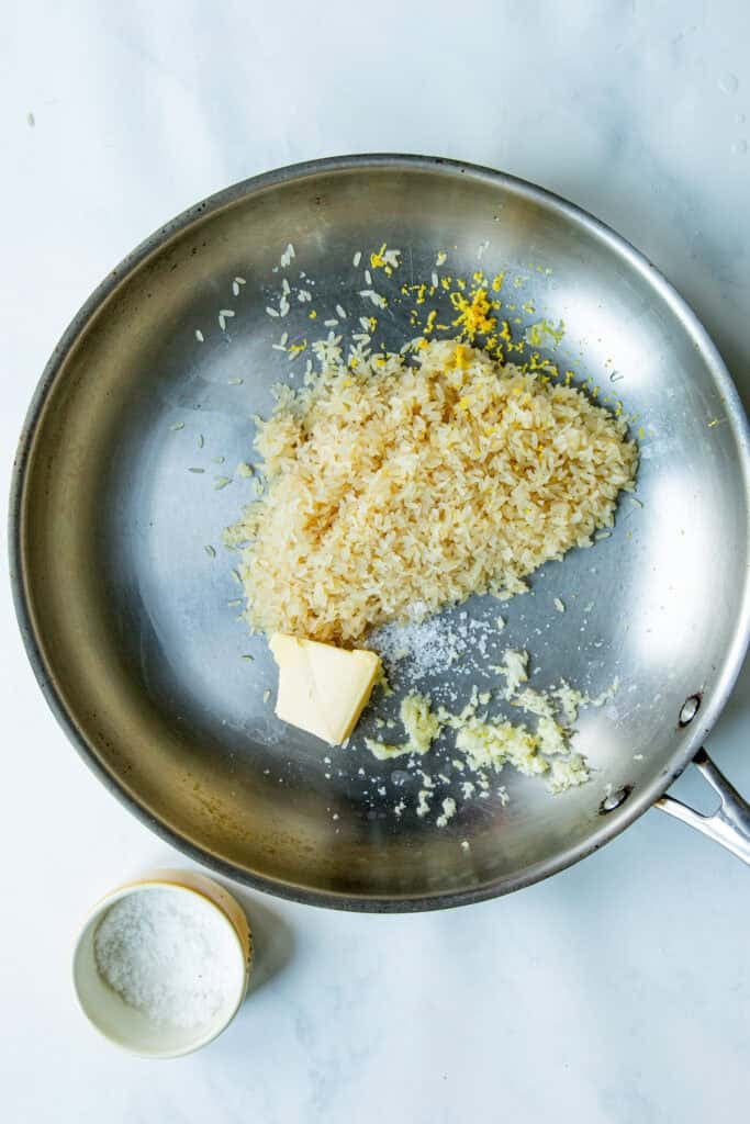 A stainless steel skillet with uncooked rice, a pat of butter, grated lemon zest, minced garlic, and salt inside—ready for cooking creamy rice. A small bowl of salt sits beside the pan on a white surface.