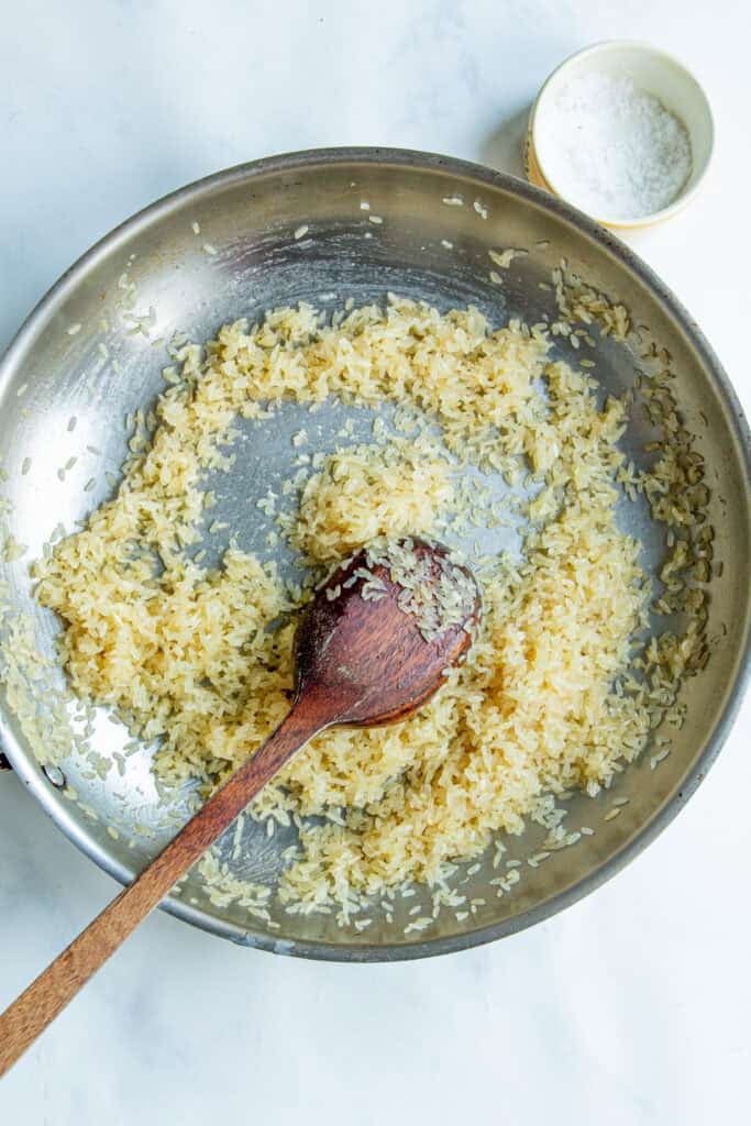 A metal skillet with uncooked rice being stirred by a wooden spoon, ready to become creamy rice, with a small bowl of salt nearby on a white surface.