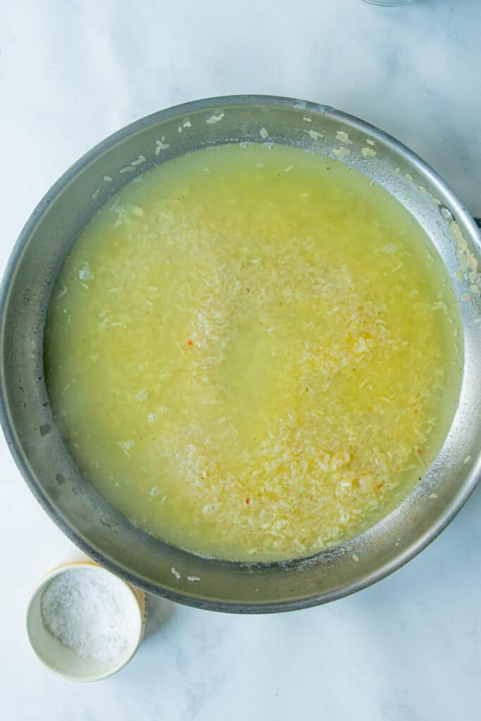 A large metal pan filled with creamy rice cooking in yellowish liquid sits on a white surface. Next to it is a small bowl containing white granulated salt.