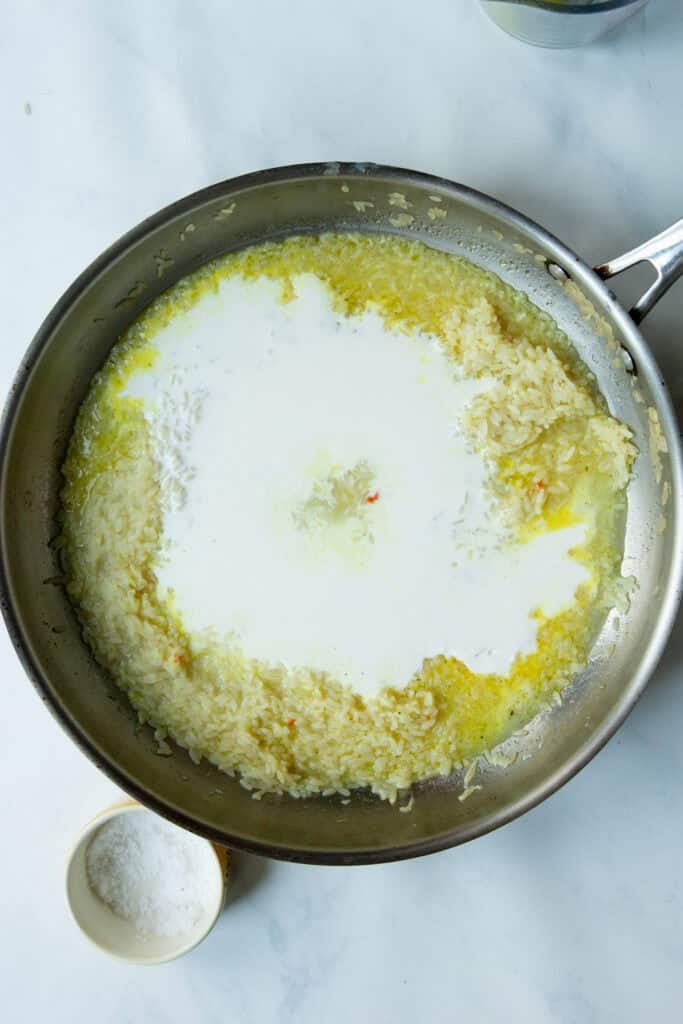 A large skillet on a white surface contains creamy rice with a pool of cream or milk in the center. A small bowl of salt sits nearby.