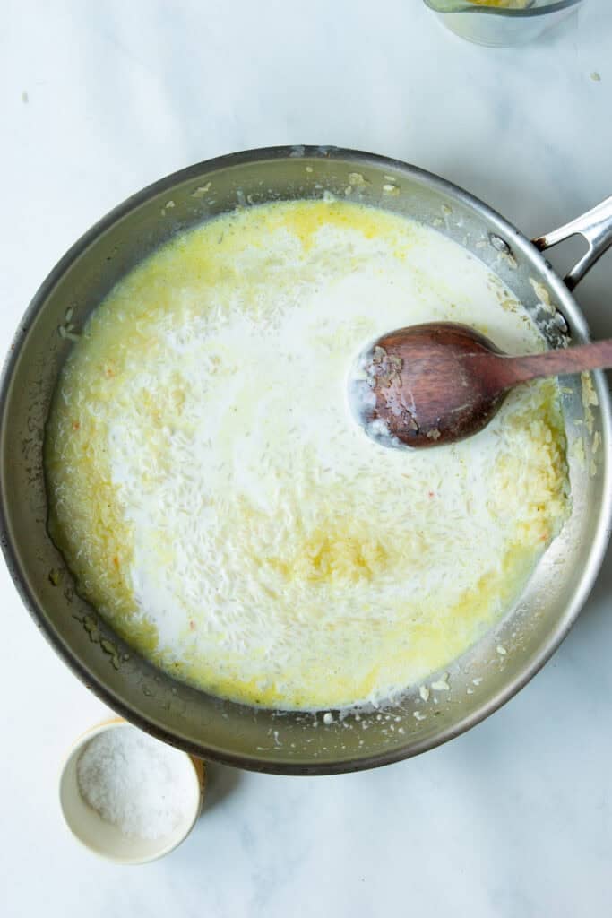 A metal skillet with creamy rice, milk, and melted butter being stirred with a wooden spoon sits on a white surface. A small bowl of coarse salt is nearby.