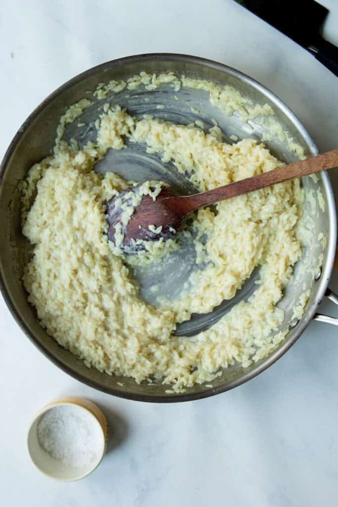 A pan of creamy rice risotto is being stirred with a wooden spoon on a white surface. A small bowl of salt sits nearby.