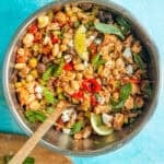 A dense bean salad in a metal bowl with chickpeas, vegetables, fresh basil leaves, lemon wedges, and a wooden spoon, set on a bright blue background.