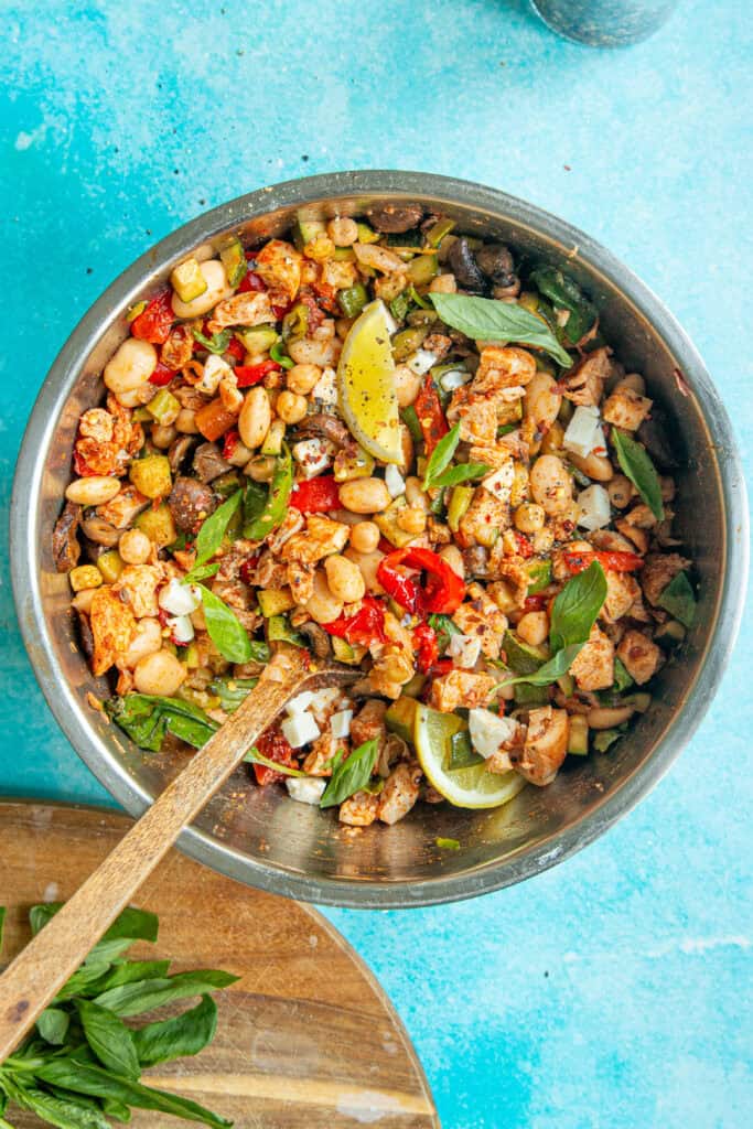 A dense bean salad in a metal bowl with chickpeas, vegetables, fresh basil leaves, lemon wedges, and a wooden spoon, set on a bright blue background.