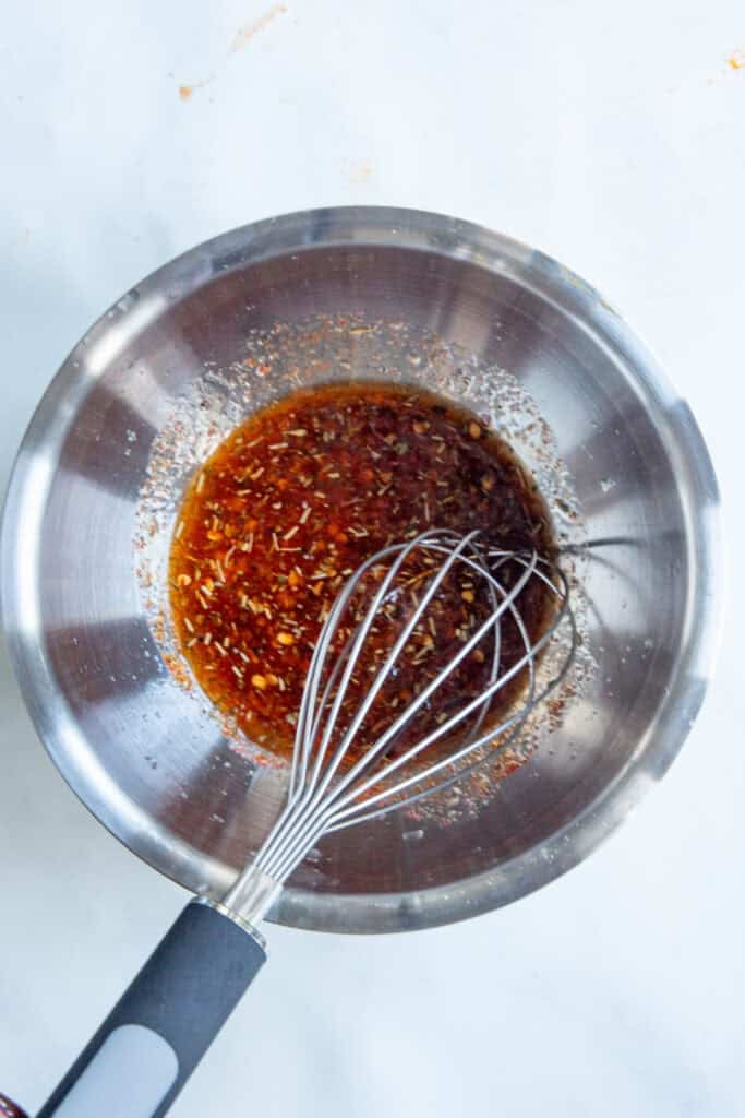 A metal mixing bowl containing a reddish-brown liquid with visible herbs and spices—perfect as a dressing for a dense bean salad—sits on a white surface with a metal whisk resting inside.