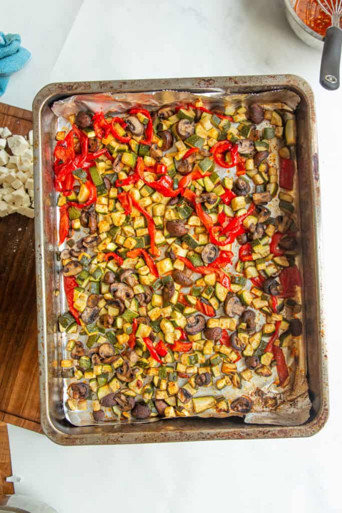A baking sheet filled with roasted vegetables, including red bell peppers, mushrooms, zucchini, and eggplant, sits on a white countertop beside chopped garlic and a wooden cutting board—perfect for pairing with a dense bean salad.