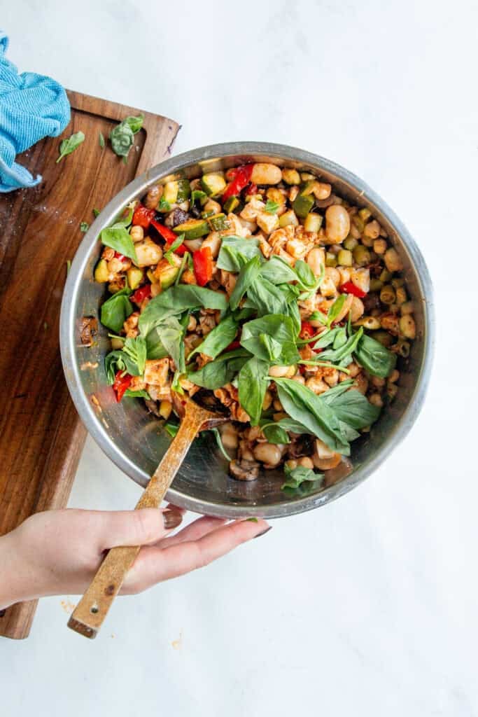 A hand holds a metal bowl filled with a dense bean salad, packed with zucchini, mushrooms, red peppers, white beans, and fresh basil leaves. A wooden spoon rests in the bowl, and a wooden cutting board is nearby.