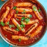 A skillet filled with devilled sausages in a rich tomato sauce, topped with fresh parsley, sits on a blue surface. A bottle and a bunch of parsley are nearby.
