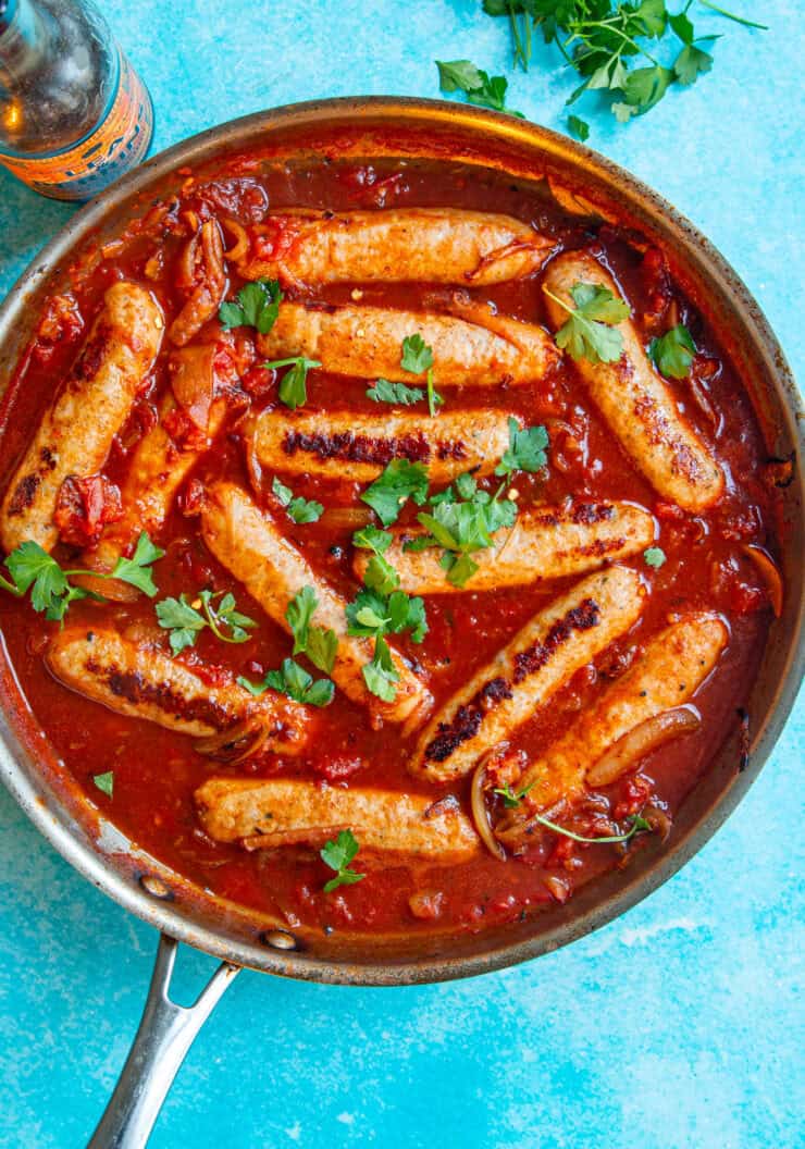 A skillet filled with devilled sausages in a rich tomato sauce, topped with fresh parsley, sits on a blue surface. A bottle and a bunch of parsley are nearby.
