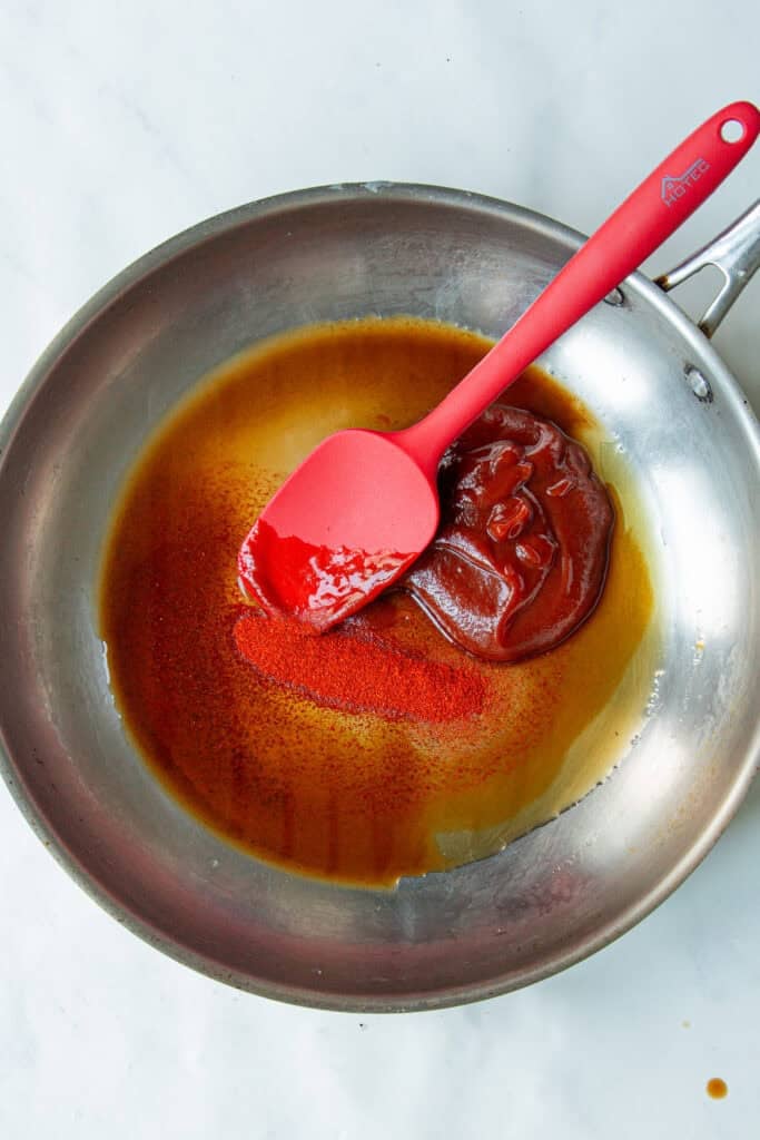 A stainless steel pan with a red silicone spatula stirring a mixture of low sugar BBQ sauce and brown liquid, possibly oil or vinegar, on a white marble surface.