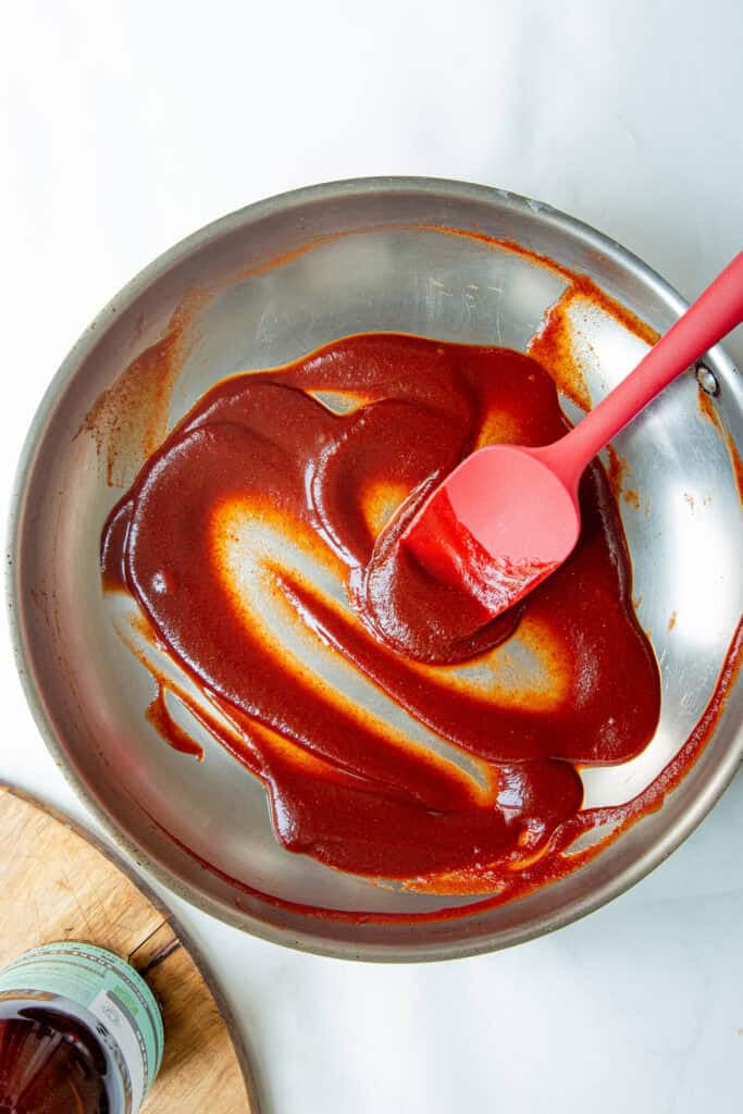 A red spatula stirs a thick, reddish-brown low sugar bbq sauce in a stainless steel pan on a white surface. A bottle sits partially visible on a wooden cutting board nearby.