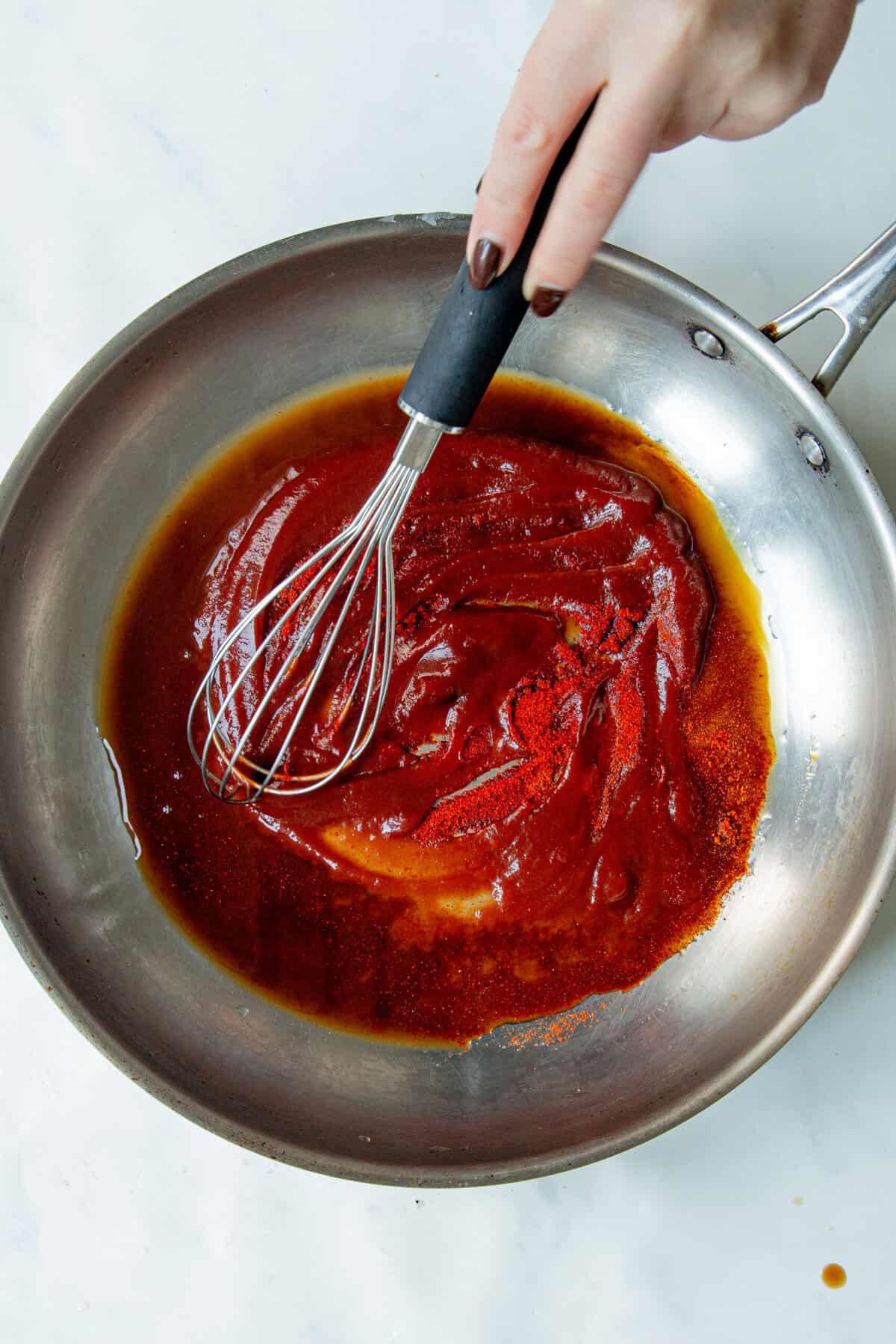 A hand holding a whisk stirs a thick, reddish-brown low sugar BBQ sauce in a stainless steel pan on a white surface.