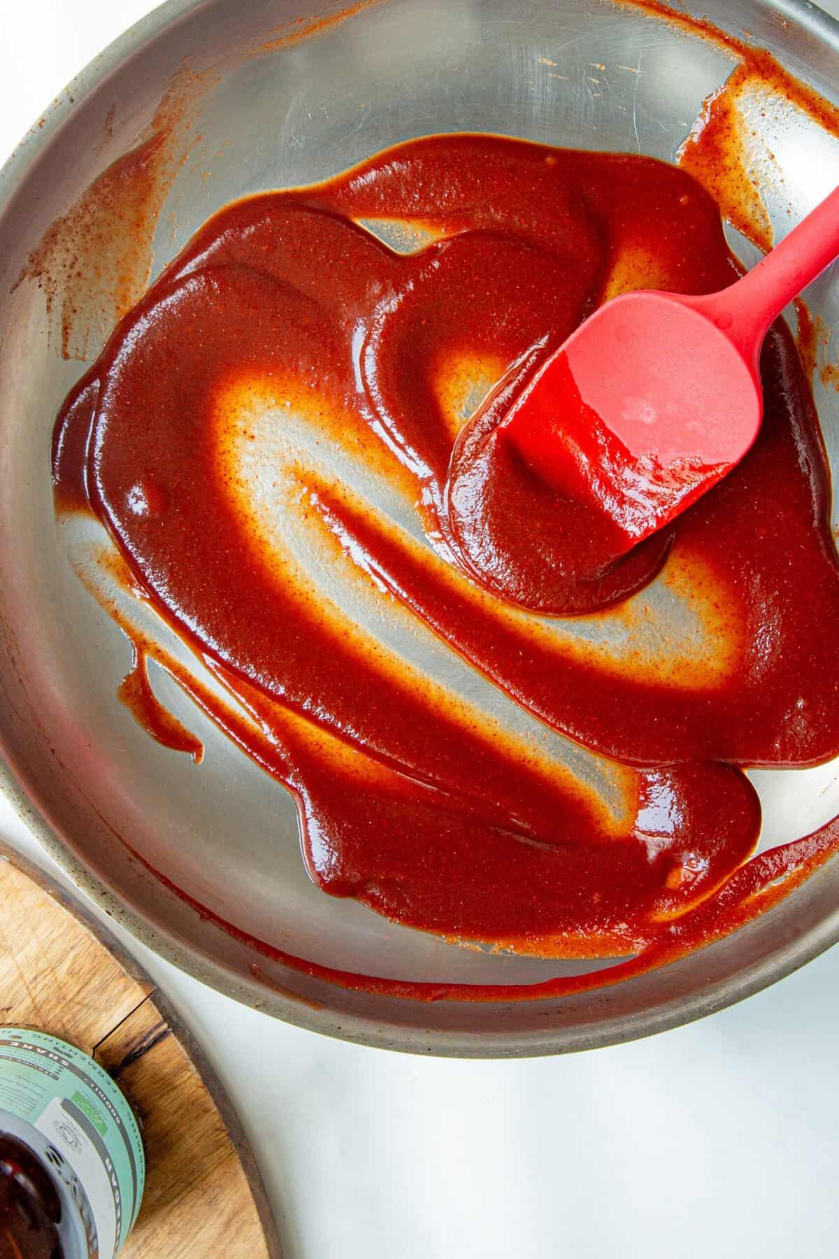 A red spatula spreading thick, glossy low sugar BBQ sauce in a metal mixing bowl; a jar is partially visible in the lower left corner of the image.