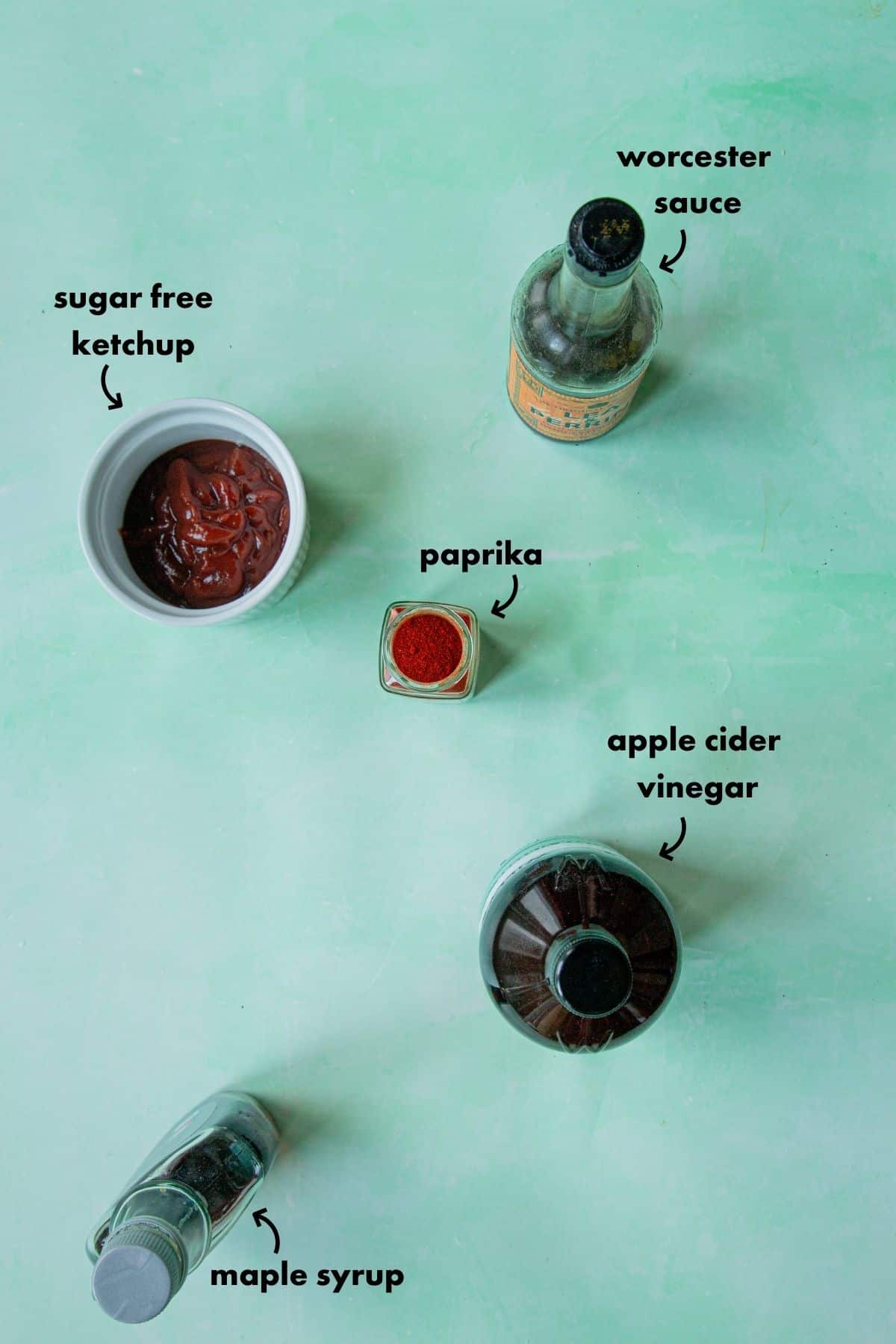 Flat lay of five low sugar BBQ sauce ingredients on a green background: sugar free ketchup in a white bowl, paprika, Worcester sauce, apple cider vinegar, and maple syrup. Each item is clearly labeled in black text.