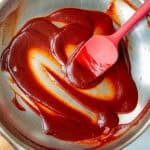 A red spatula spreading thick, dark red low sugar bbq sauce in a stainless steel mixing bowl.