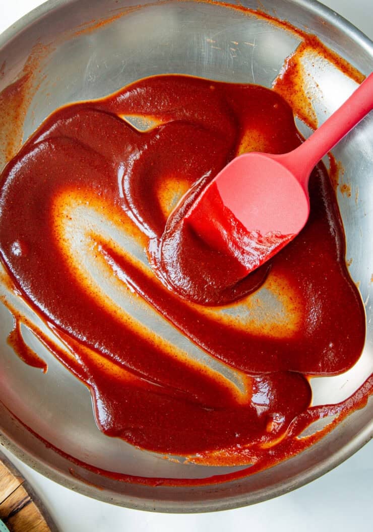 A red spatula spreading thick, dark red low sugar bbq sauce in a stainless steel mixing bowl.