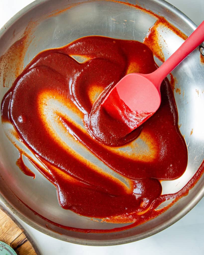 A red spatula spreading thick, dark red low sugar bbq sauce in a stainless steel mixing bowl.