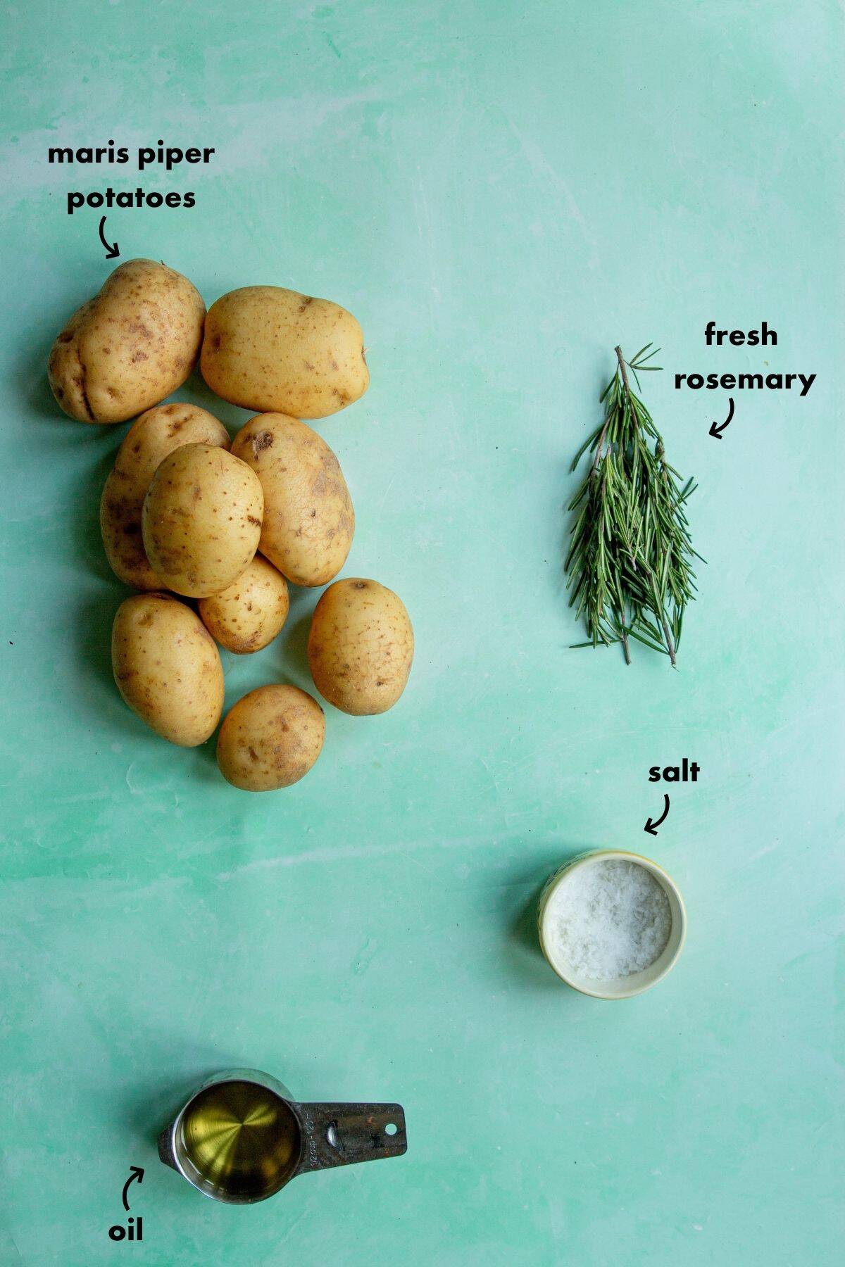 Overhead view of Maris Piper potatoes, a bunch of fresh rosemary, a small cup of salt, and a metal measuring cup with oil arranged on a light green surface. Labels identify each ingredient.