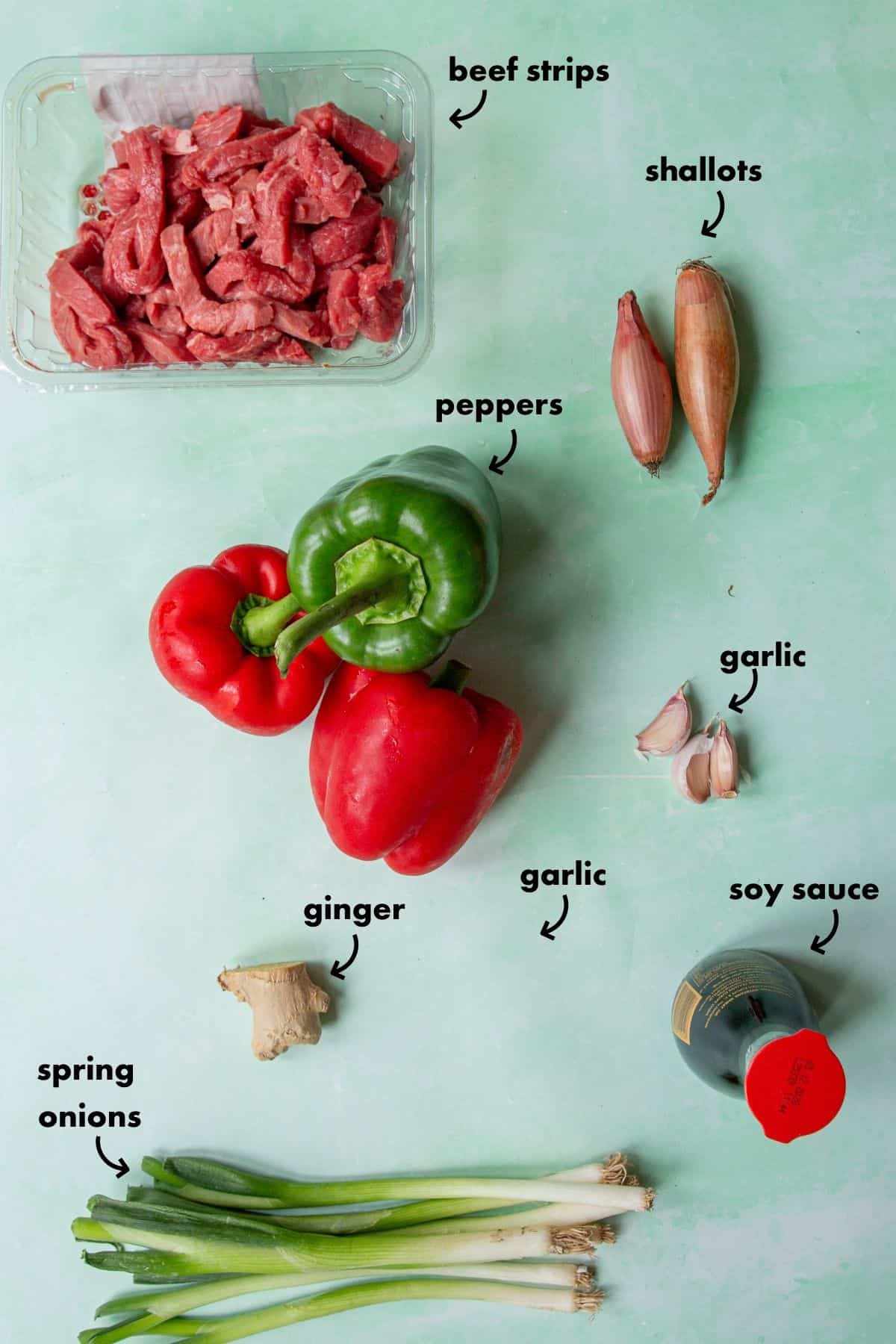 A flat lay of beef strips, two shallots, two red bell peppers, one green bell pepper, roasted corn, two garlic cloves, a piece of ginger, spring onions, and a bottle of soy sauce on a light green background.